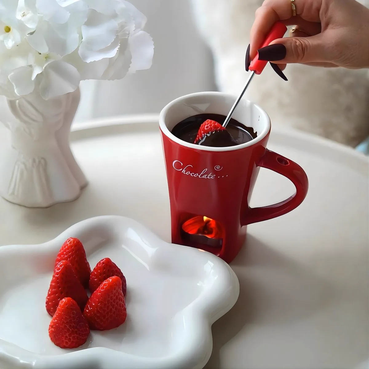 A hand dipping a strawberry into dark chocolate in the red mug, with a plate of strawberries in front.