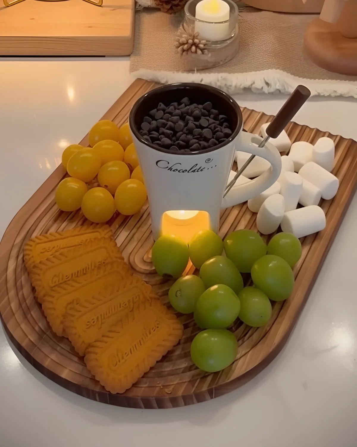 Sharing board with the white mug in the middle, chocolate chips inside, surrounded by marshmallows, grapes, tomatoes and biscuits.